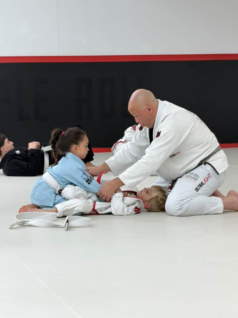 Professor Paraíba kneeling on the mat, showing a young student a technique during kids class